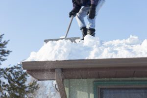 A man is shoveling snow off of a house roof after a blizzard.