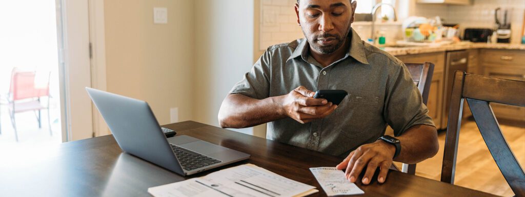 Man in kitchen sitting down in front of his laptop taking photos of his receipts